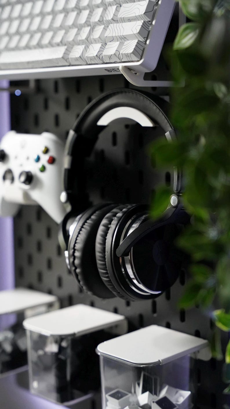 Puede incluir: Auriculares negros de diadema con diadema plateada colgados en una pared de tablero perforado. Tambi&eacute;n se puede ver un mando de videojuegos blanco y dos recipientes de pl&aacute;stico transparente con tapas blancas.