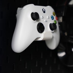 Può includere: Un controller Xbox bianco con pulsanti neri e un pulsante verde, giallo e rosso sul davanti. Il controller è montato su una parete di pannelli forati neri.