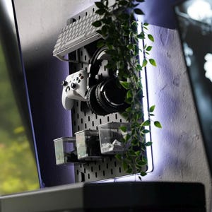 Puede incluir: Un tablero de clavijas blanco con un teclado blanco, unos auriculares negros, un mando de videojuegos blanco y recipientes de pl&aacute;stico transparentes. El tablero de clavijas est&aacute; montado en una pared gris con una planta colgando delante. La pared est&aacute; iluminada con una tira LED p&uacute;rpura.