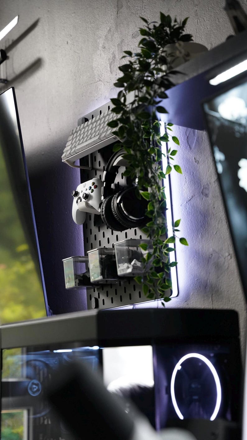 Puede incluir: Un tablero de clavijas blanco con un teclado blanco, unos auriculares negros, un mando de videojuegos blanco y recipientes de pl&aacute;stico transparentes. El tablero de clavijas est&aacute; montado en una pared gris con una planta colgando delante. La pared est&aacute; iluminada con una tira LED p&uacute;rpura.