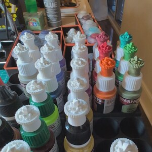 May include: A collection of small paint bottles with white, crystal-shaped bottle stoppers. The bottles are arranged in a grid pattern and are filled with various colors of paint, including green, red, orange, and black.