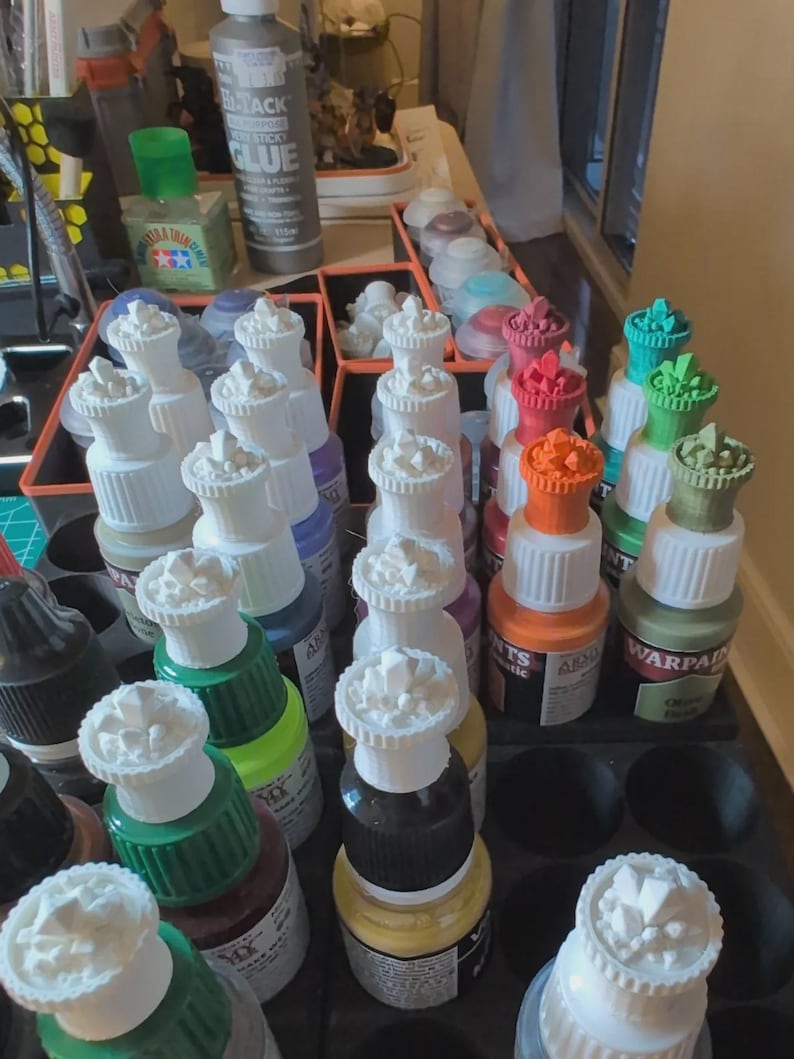 May include: A collection of small paint bottles with white, crystal-shaped bottle stoppers. The bottles are arranged in a grid pattern and are filled with various colors of paint, including green, red, orange, and black.