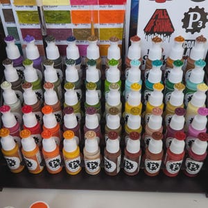 May include: A collection of 60 small bottles of paint, arranged in rows on a black shelf. The bottles are labeled with the letter "P" and a number. The paint colors are a variety of shades of brown, red, orange, yellow, green, blue, and purple.