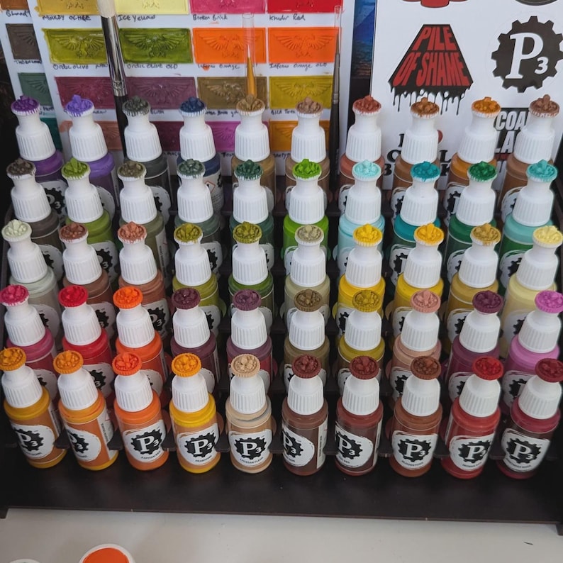 May include: A collection of 60 small bottles of paint, arranged in rows on a black shelf. The bottles are labeled with the letter "P" and a number. The paint colors are a variety of shades of brown, red, orange, yellow, green, blue, and purple.