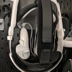 Puede incluir: Un auricular de realidad virtual blanco con una correa negra y un mando blanco colgando de un tablero de clavijas. La correa tiene la palabra "Zyber" impresa. El auricular tiene el texto "Pay Attention to the Light" impreso en la parte inferior.