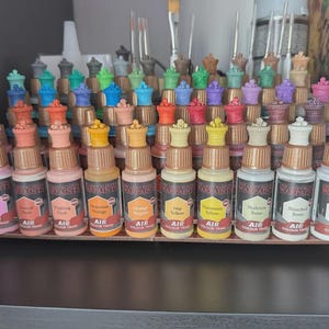 May include: A set of 18 small bottles of acrylic paint, arranged in a row on a wooden shelf. The bottles are labeled with the paint color, such as "Sky Blue", "Bleached Bone", and "Orange Magenta". The bottles are all the same size and shape, and they have a black and red label with the words "Warpaint Air Colour" printed on them.