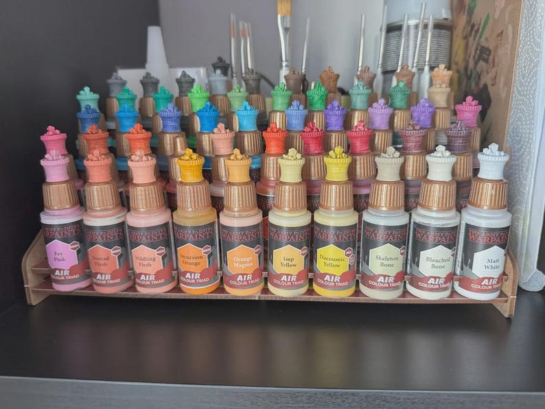 May include: A set of 18 small bottles of acrylic paint, arranged in a row on a wooden shelf. The bottles are labeled with the paint color, such as "Sky Blue", "Bleached Bone", and "Orange Magenta". The bottles are all the same size and shape, and they have a black and red label with the words "Warpaint Air Colour" printed on them.