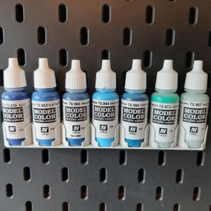 Könnte beinhalten: Acht kleine Flaschen Acrylfarbe in verschiedenen Blautönen und Türkis. Die Flaschen sind mit dem Markennamen "Model Color" und der Farbnummer beschriftet. Die Flaschen sind in einer Reihe auf einem weißen Regal angeordnet.