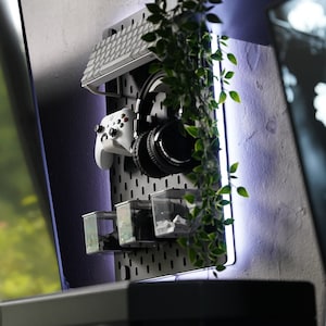 May include: A white gaming controller and black headphones are hanging on a pegboard with a white keyboard and small containers. A green plant hangs down from the top of the pegboard.
