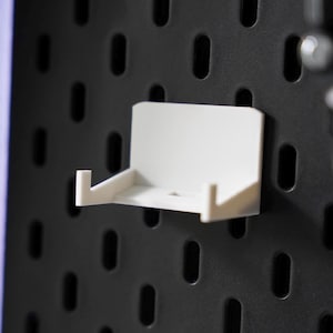 Puede incluir: Estante de pared de pl&aacute;stico blanco con un peque&ntilde;o borde en la parte delantera. El estante est&aacute; montado en una pared de clavijas negra.