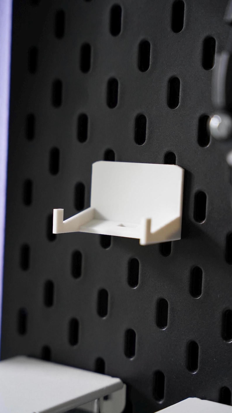 Puede incluir: Estante de pared de pl&aacute;stico blanco con un peque&ntilde;o borde en la parte delantera. El estante est&aacute; montado en una pared de clavijas negra.