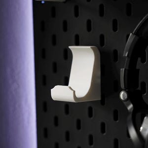 Puede incluir: Soporte para auriculares de pl&aacute;stico blanco montado en una pared de tablero de clavijas negro. El soporte tiene forma de gancho con la parte superior curva y la parte inferior plana.