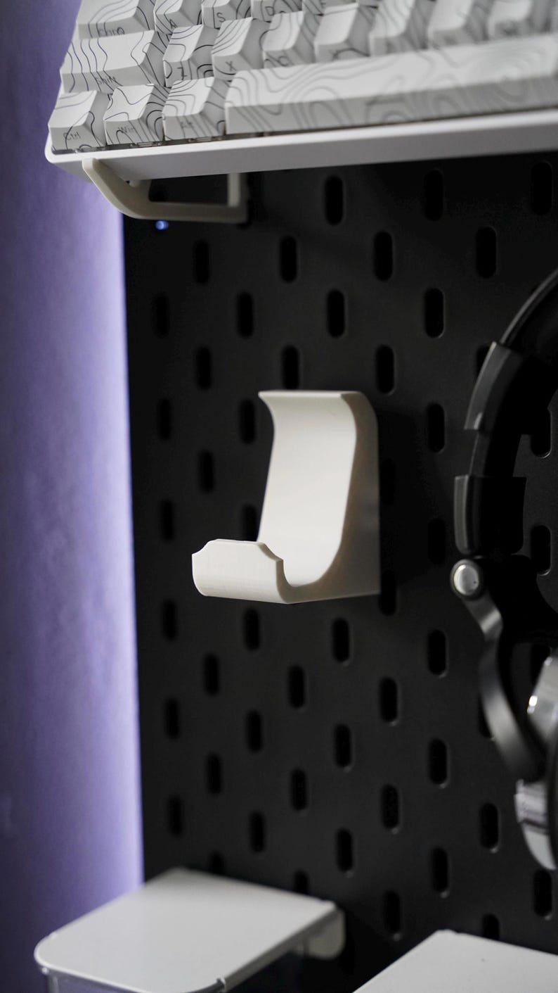 Puede incluir: Soporte para auriculares de pl&aacute;stico blanco montado en una pared de tablero de clavijas negro. El soporte tiene forma de gancho con la parte superior curva y la parte inferior plana.