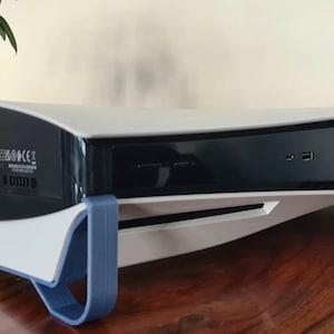 Peut inclure: Une console PlayStation 5 blanche avec un support bleu. La console a un panneau frontal noir avec des ports et un panneau supérieur blanc. La console est posée sur une surface en bois.