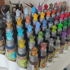 May include: A collection of small bottles of paint, organized in a white plastic rack. The bottles are arranged in rows and columns, and each bottle has a different color of paint. The bottles are labeled with the words "Speed Paint" and the color of the paint.