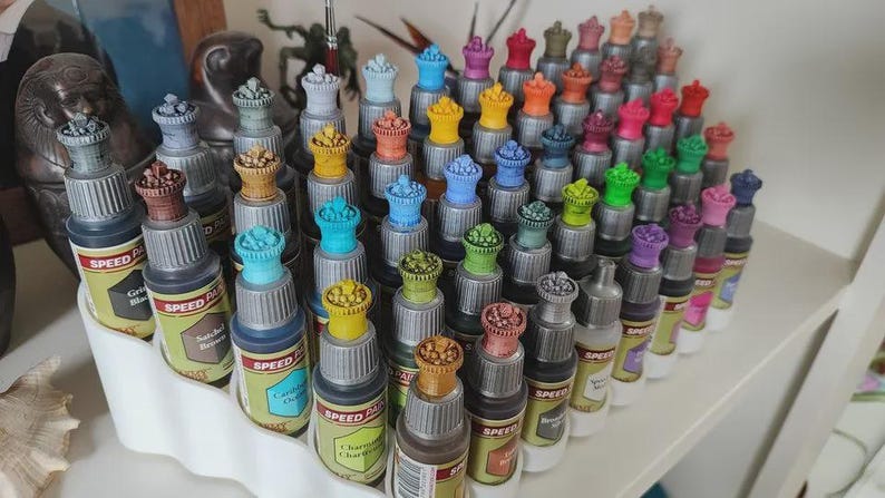 May include: A collection of small bottles of paint, organized in a white plastic rack. The bottles are arranged in rows and columns, and each bottle has a different color of paint. The bottles are labeled with the words "Speed Paint" and the color of the paint.