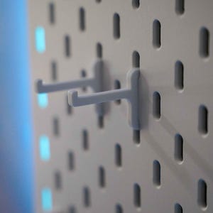 Könnte beinhalten: Zwei weiße Kunststoff-Pegboard-Haken sind an einem weißen Pegboard mit blauem Hintergrund befestigt.