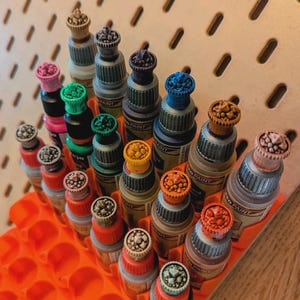 May include: A collection of small paint bottles with colorful, textured caps arranged in an orange egg carton. The bottles are labeled "Model Paint" and have a variety of colors, including red, blue, green, and brown.