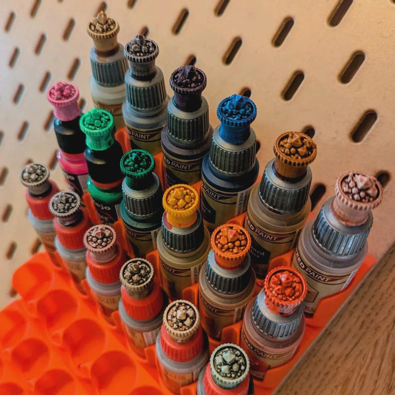 May include: A collection of small paint bottles with colorful, textured caps arranged in an orange egg carton. The bottles are labeled "Model Paint" and have a variety of colors, including red, blue, green, and brown.