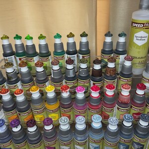 May include: A collection of small bottles of acrylic paint, organized on a shelf. The bottles are labeled "Speed Paint" and feature a variety of colors, including red, green, blue, yellow, and black. The bottles are arranged in rows, with each row containing a different color.