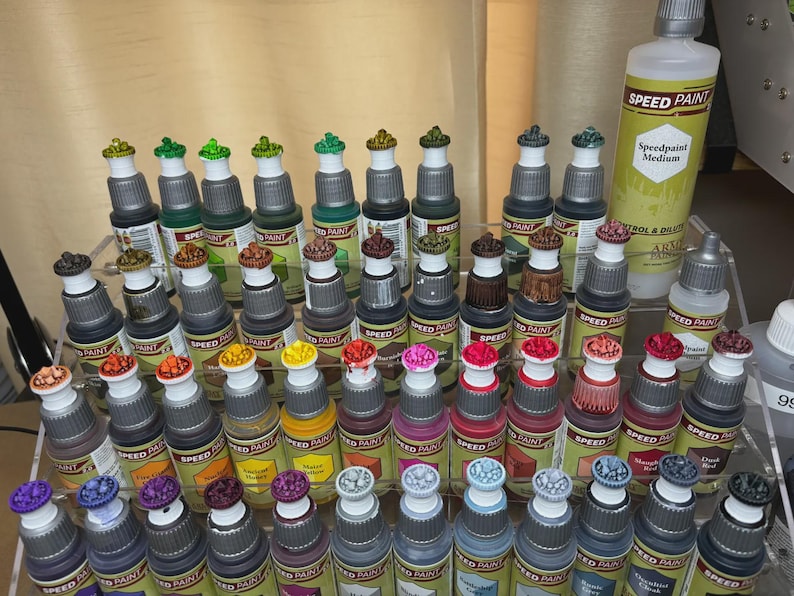 May include: A collection of small bottles of acrylic paint, organized on a shelf. The bottles are labeled "Speed Paint" and feature a variety of colors, including red, green, blue, yellow, and black. The bottles are arranged in rows, with each row containing a different color.