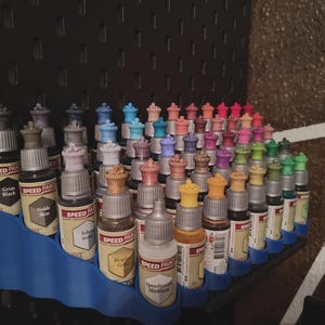 May include: A collection of small bottles of paint organized in a blue plastic rack. The bottles are arranged in rows and are labeled with different colors and names, such as "Grim Black", "Noble Skin", "Speedpaint Medium".