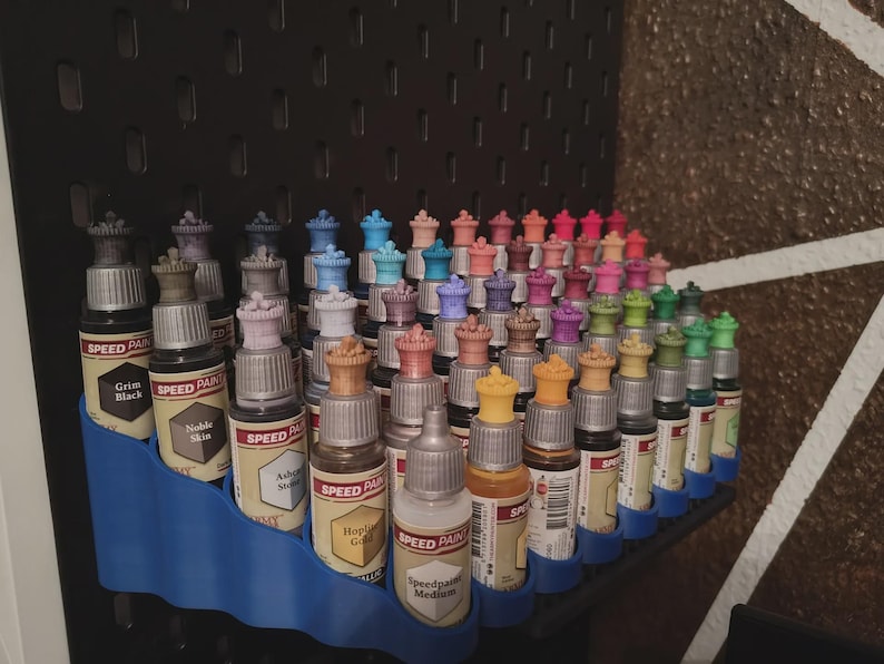 May include: A collection of small bottles of paint organized in a blue plastic rack. The bottles are arranged in rows and are labeled with different colors and names, such as "Grim Black", "Noble Skin", "Speedpaint Medium".