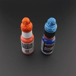 Op de afbeelding: Twee flessen verf, een oranje en een blauwe. De flessen hebben decoratieve doppen met een textuurontwerp. De oranje fles is ge&euml;tiketteerd met "Model Color" en de blauwe fles is ge&euml;tiketteerd met "Vallejo Xpress Color".