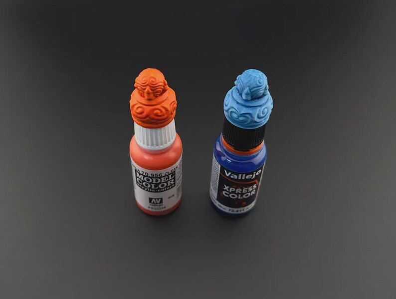 Op de afbeelding: Twee flessen verf, een oranje en een blauwe. De flessen hebben decoratieve doppen met een textuurontwerp. De oranje fles is ge&euml;tiketteerd met "Model Color" en de blauwe fles is ge&euml;tiketteerd met "Vallejo Xpress Color".