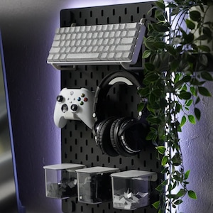 May include: A black pegboard with white LED lights around the perimeter. The pegboard has a white keyboard, a white video game controller, black headphones, and three clear plastic containers with white paper inside.