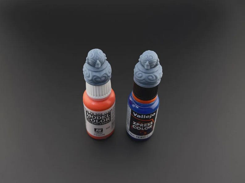Op de afbeelding: Twee flessen verf met decoratieve doppen. De fles links is oranje en heeft "Model Color" op het etiket staan. De fles rechts is blauw en heeft "Vallejo Xpress Color" op het etiket staan.