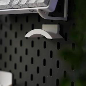 Puede incluir: Un teclado blanco con un patr&oacute;n topogr&aacute;fico en las teclas est&aacute; montado en una pared de tablero de clavijas negro. Un gancho de pl&aacute;stico blanco tambi&eacute;n est&aacute; montado en la pared.