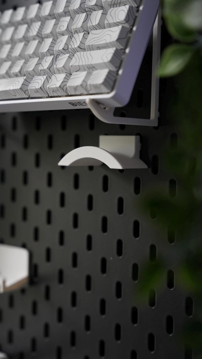 Puede incluir: Un teclado blanco con un patr&oacute;n topogr&aacute;fico en las teclas est&aacute; montado en una pared de tablero de clavijas negro. Un gancho de pl&aacute;stico blanco tambi&eacute;n est&aacute; montado en la pared.