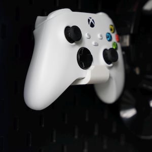 Puede incluir: Un mando Xbox blanco con botones negros y un bot&oacute;n verde, amarillo y rojo en la parte delantera. El mando est&aacute; montado en una placa perforada negra.