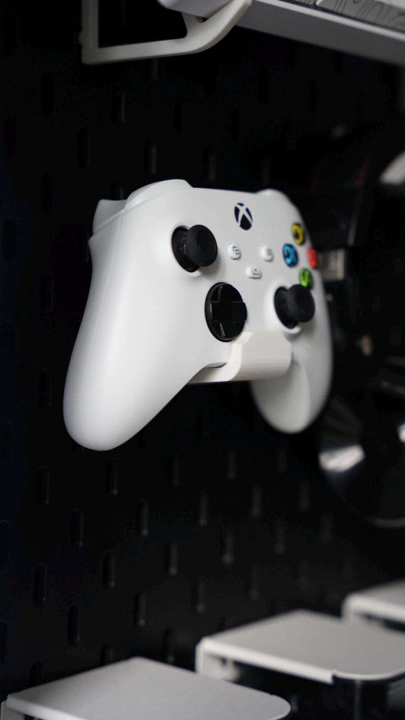 Puede incluir: Un mando Xbox blanco con botones negros y un bot&oacute;n verde, amarillo y rojo en la parte delantera. El mando est&aacute; montado en una placa perforada negra.