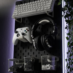 May include: A black pegboard with a white keyboard, black headphones, a white video game controller, and three clear plastic containers with black and white objects inside. The pegboard has a white light around the edges.