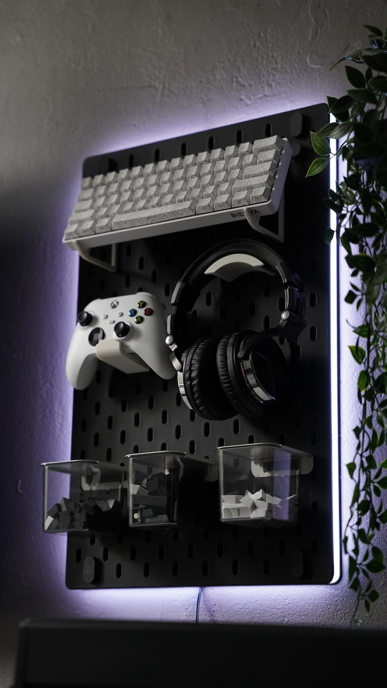 May include: A black pegboard with a white keyboard, black headphones, a white video game controller, and three clear plastic containers with black and white objects inside. The pegboard has a white light around the edges.