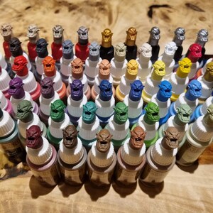 Puede incluir: Una colecci&oacute;n de 40 peque&ntilde;os frascos de pintura con tapas de color en forma de duende. Los frascos est&aacute;n dispuestos en filas sobre una superficie de madera.