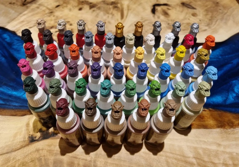 Puede incluir: Una colecci&oacute;n de 40 peque&ntilde;os frascos de pintura con tapas de color en forma de duende. Los frascos est&aacute;n dispuestos en filas sobre una superficie de madera.