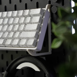 May include: A white keyboard with a wood grain pattern is mounted on a black pegboard wall. A pair of black headphones are hanging below the keyboard.