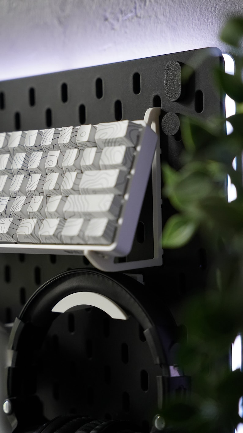 May include: A white keyboard with a wood grain pattern is mounted on a black pegboard wall. A pair of black headphones are hanging below the keyboard.