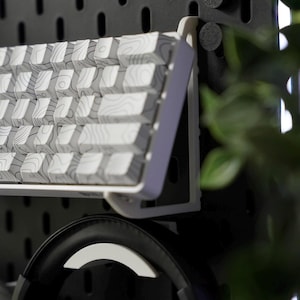 Puede incluir: Un teclado blanco con un patr&oacute;n de veta de madera est&aacute; montado en una pared de tablero de clavijas negro. Un par de auriculares negros cuelgan debajo del teclado.