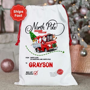 May include: White Christmas gift bag with red drawstring closure. Features a red truck driven by Santa Claus, delivering gifts. Text includes "North Pole Express Delivery" and "Grayson".