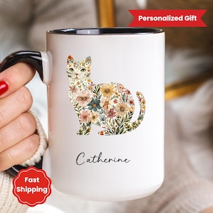 May include: White ceramic mug with a black handle and rim, featuring a floral cat design. The cat is composed of various flowers in shades of pink, yellow, and blue. The name "Catherine" is written below the cat in a script font.