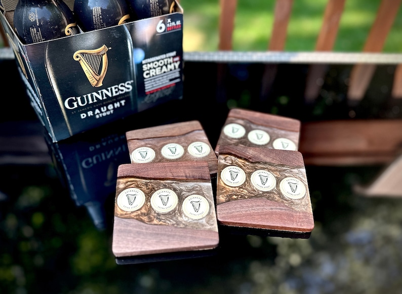 Pu&ograve; includere: Cinque sottobicchieri in legno marrone con un design in resina epossidica bianca e dorata. Ogni sottobicchiere presenta un motivo circolare con il logo Guinness e un'arpa.