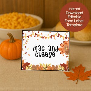 May include: A white rectangular food label with the text "mac and cheese" in a whimsical font. The label has a turkey illustration and autumn-themed decorations. A small orange pumpkin and a fall leaf are in the foreground.
