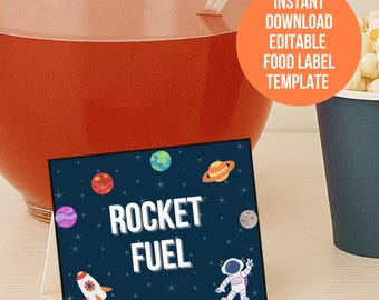 以下が含まれることがあります： 白い「ROCKET FUEL」の文字が入った、ネイビーブルーの食品ラベル。周りには漫画の惑星、ロケット、宇宙飛行士が描かれています。ラベルは、赤い液体が入ったガラスのボウルと青い紙コップの隣にあります。