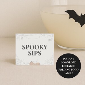 Op de afbeelding: Een rechthoekige kaart met de tekst "SPOOKY SIPS" in grijs, met een spinnenwebontwerp en kleine pompoenen. Een zwarte cirkel met witte tekst luidt "INSTANT DOWNLOAD EDITABLE FOLDING FOOD LABELS". Een glas met een vleermuissilhouet staat op de achtergrond.