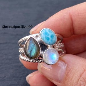 May include: A multi-band silver ring featuring three distinct gemstones: a pear-shaped iridescent labradorite, an oval sky-blue larimar, and a round translucent moonstone. The band includes a twisted silver wire detail and small clear stones.