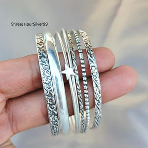 May include: A set of silver bangles, some with floral patterns and others with a smooth, polished finish. One bangle features a star charm. The bangles are stacked together, showcasing their varying designs and textures.
